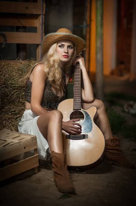 A blonde woman in a straw hat and cowboy boots, sitting on a hay bale with an acoustic guitar.